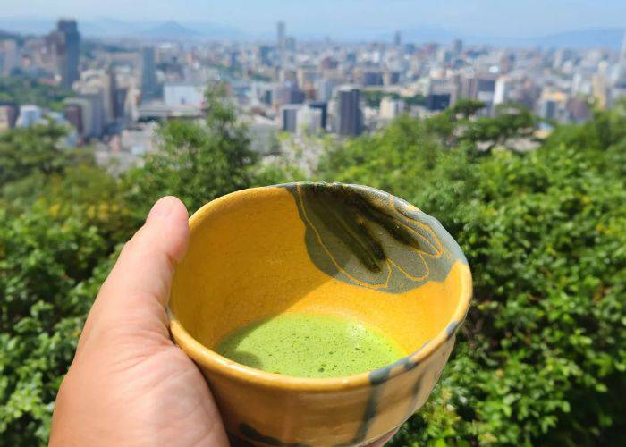 A hand holding out a matcha cup over the skyline of Hiroshima City.
